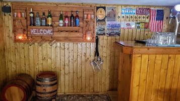 A bar area with wooden walls, a welcome sign, and various liquor bottles. There are wooden barrels and a tiled floor.