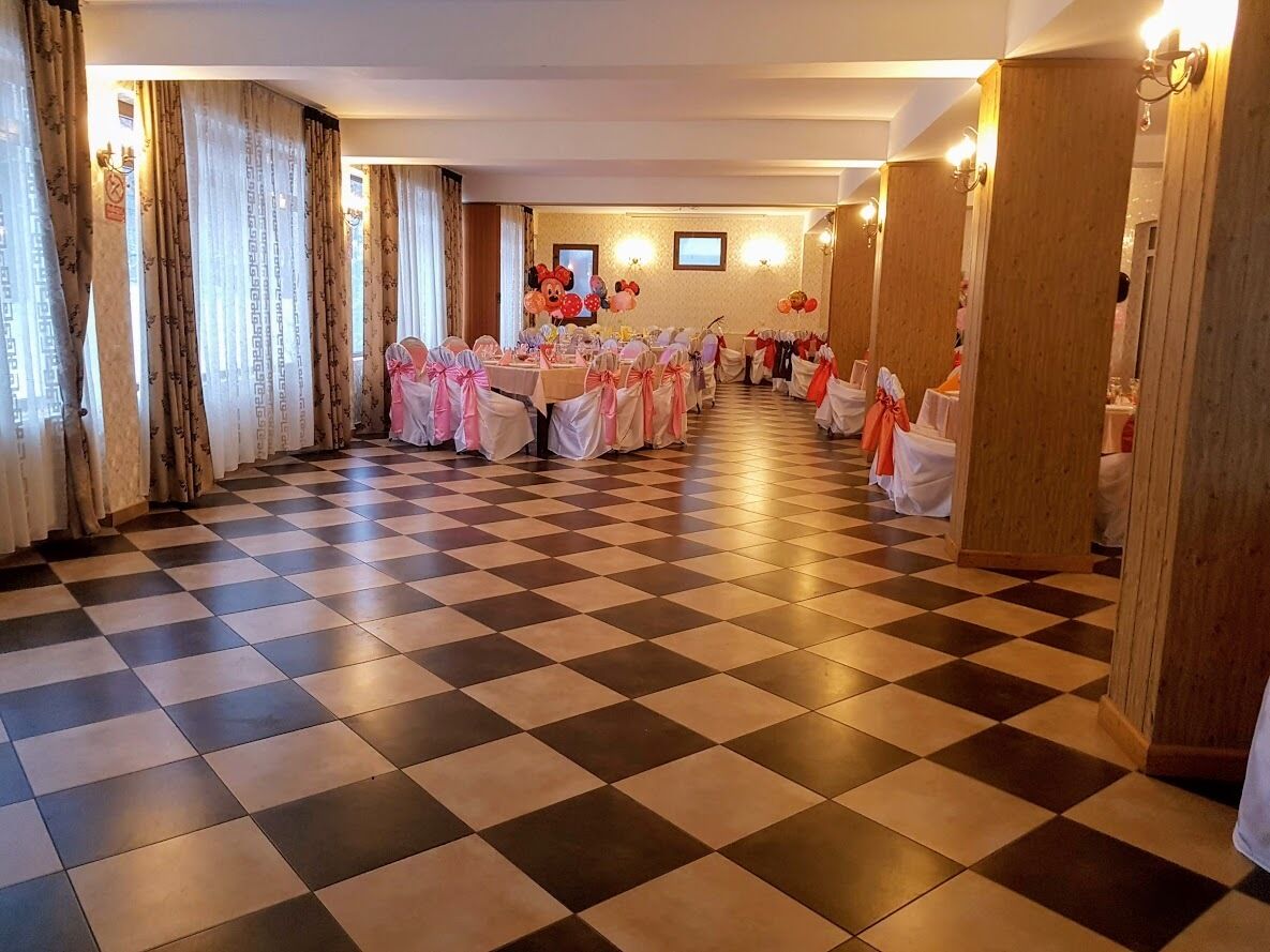 Een feestzaal met geruit vloerkleed, tafels met witte tafelkleden en roze linten, en houten lambrisering.