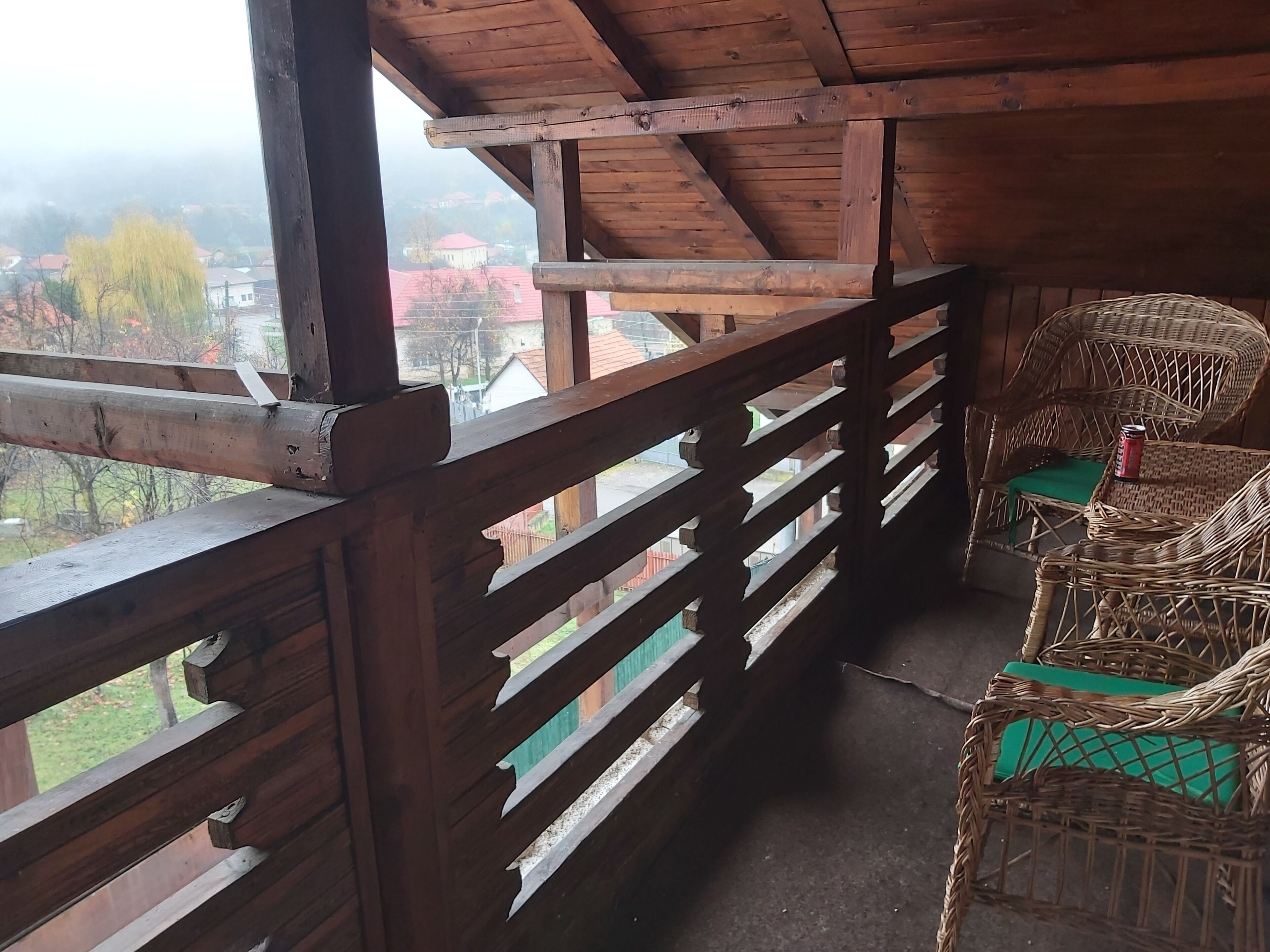 A wooden balcony with a view of buildings and trees. There are wicker chairs with green cushions.