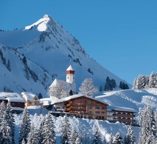 A snow-covered mountain with a church and wooden buildings in the foreground. - Boutique Hotel die Mittagspitze Superior (Damüls)