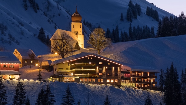 A snow-covered mountain lodge with a prominent church tower, surrounded by snow-laden trees and a clear sky. - Boutique Hotel die Mittagspitze Superior (Damüls)