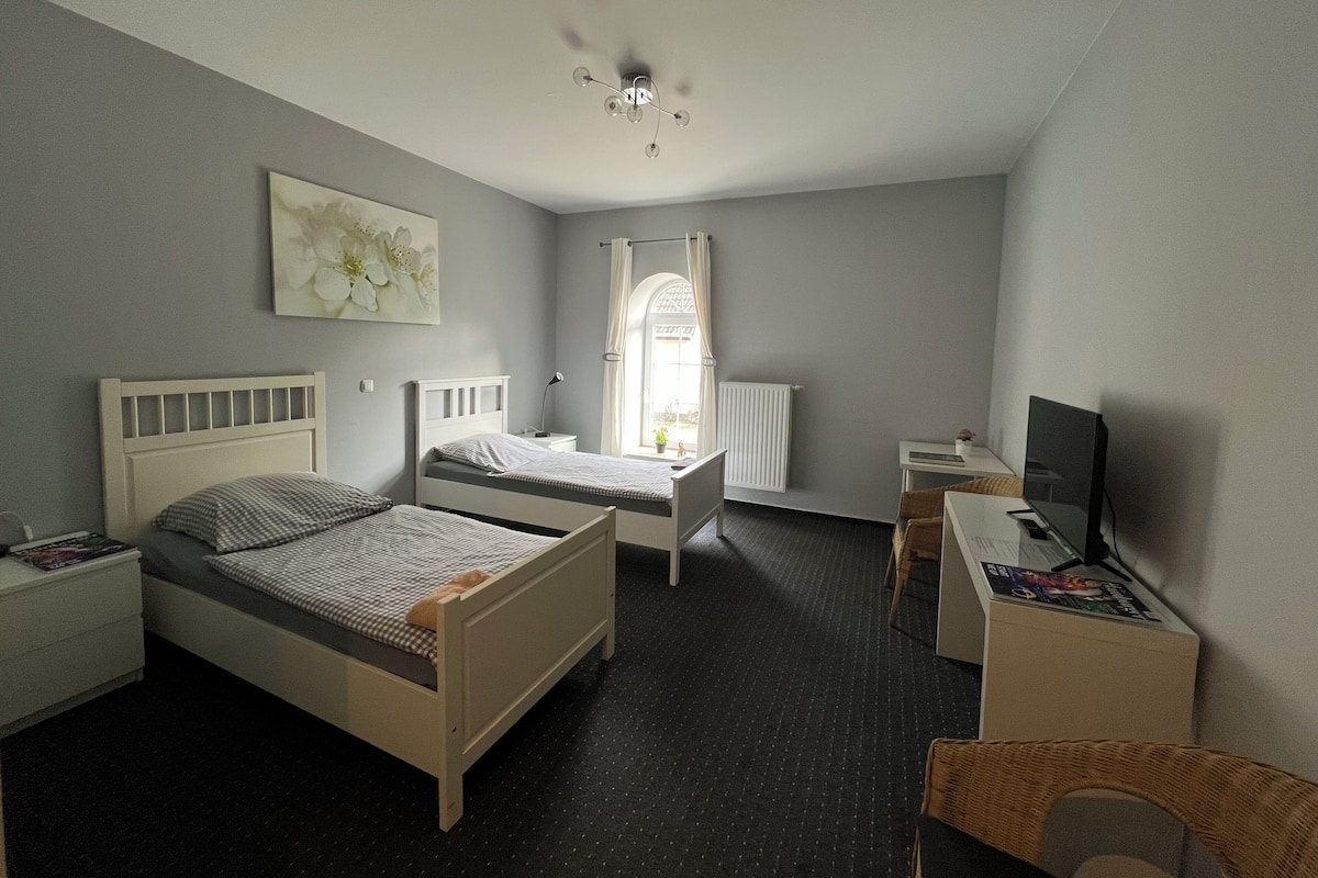 Un dormitorio con dos camas individuales, un escritorio con computadora, una silla, un radiador y una ventana con cortinas.