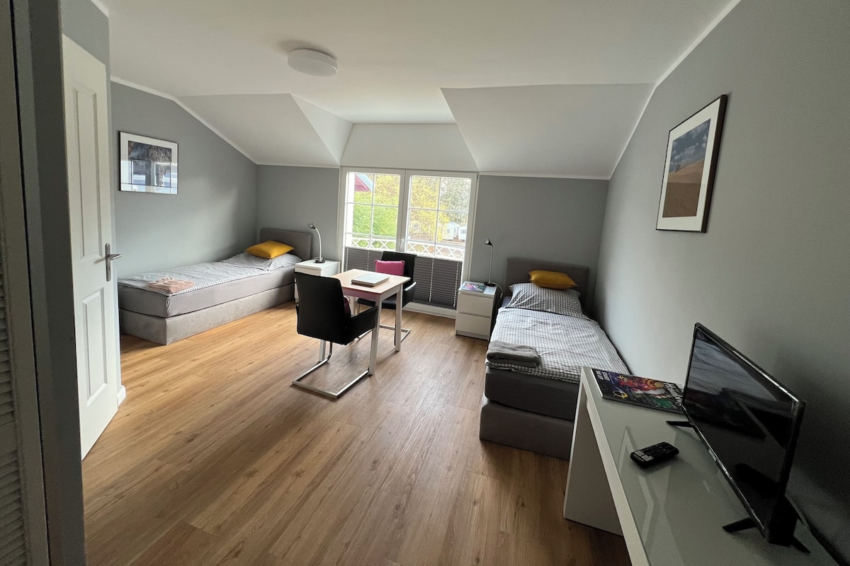 Habitación compacta con dos camas individuales, un escritorio con silla, una mesita pequeña, un televisor y una ventana con persianas.