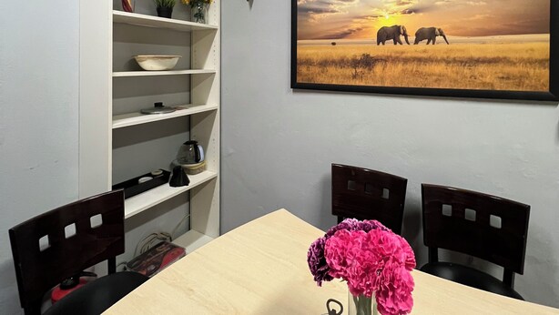 Un comedor con una mesa de madera, cuatro sillas, un florero con flores, una estantería y un cuadro con elefantes.