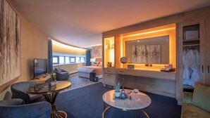 Ein modernes Hotelzimmer mit einem großen Bett, einer Sitzecke mit Sofa und Sesseln, einem kleinen Tisch mit Getränken und einem begehbaren Kleiderschrank mit Bademänteln.