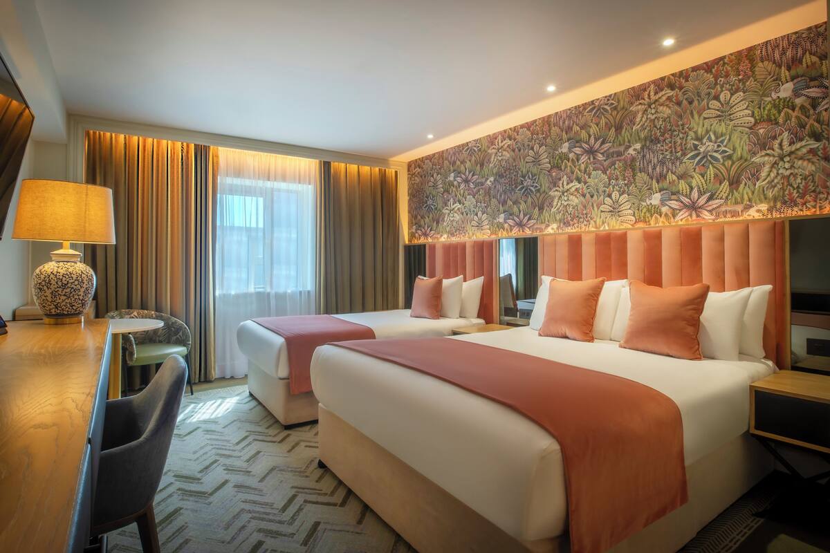 A hotel room with two beds, a desk, a chair, a TV, and a large floral wall mural.