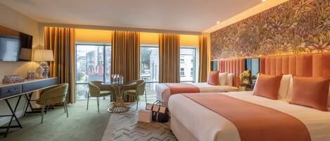 Quarto de hotel com duas camas, uma escrivaninha com abajur, uma cadeira, uma mesa com um vaso e uma janela grande com cortinas.
