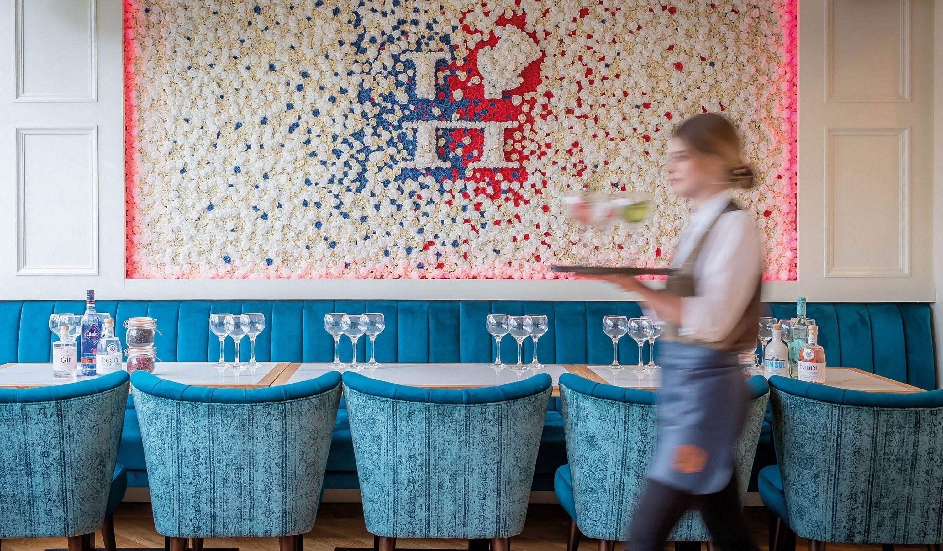 En restaurant med en væg af dekorative flasker, teal-polstrede stole og en tjener, der bærer en bakke.