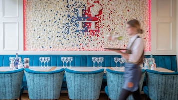 Un restaurante con una pared de botellas decorativas, sillas tapizadas en color turquesa y un camarero llevando una bandeja.