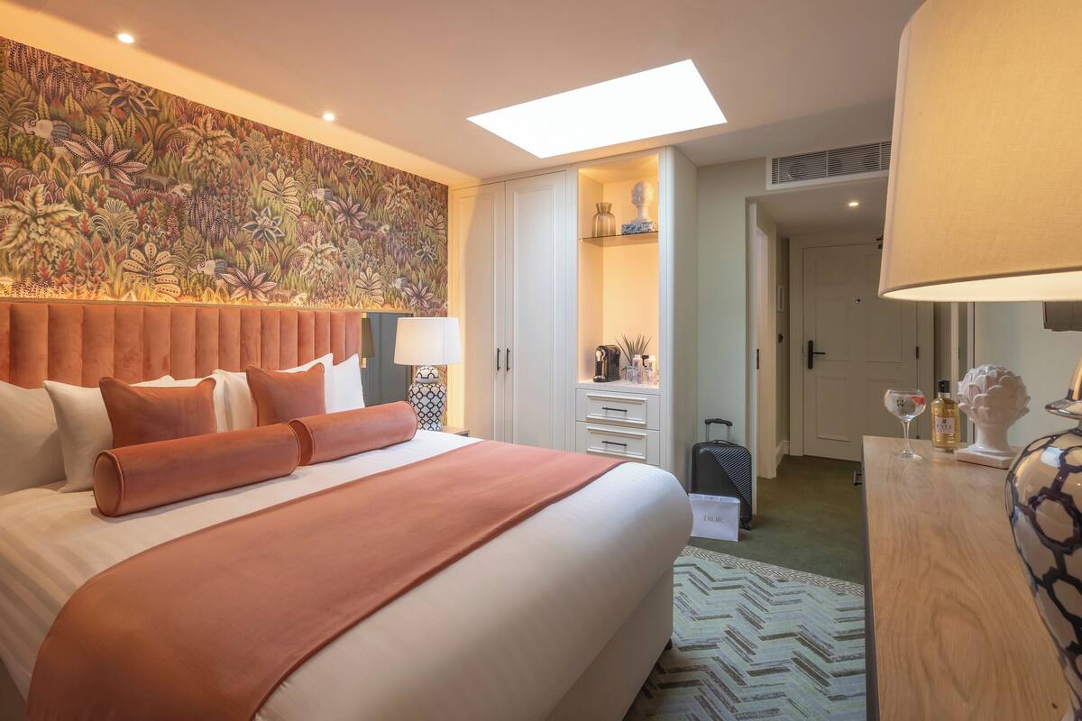 A hotel room with a large bed, a headboard with a floral tapestry, a skylight, a bedside table with a lamp, and a dresser with a vase and a bottle.