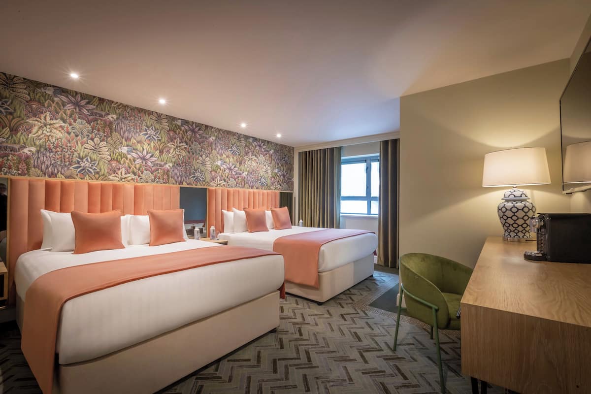 A hotel room with two beds, a desk, a chair, a patterned wall, and a window with curtains.
