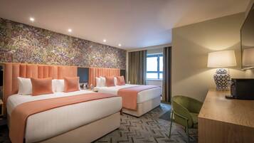 A hotel room with two beds, a desk, a chair, a patterned wall, and a window with curtains.