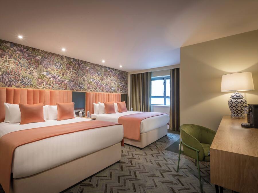 A hotel room with two beds, a desk, a chair, a patterned wall, and a window with curtains.