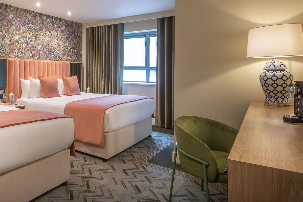 A hotel room with two beds, a green chair, a lamp, and a window with curtains.