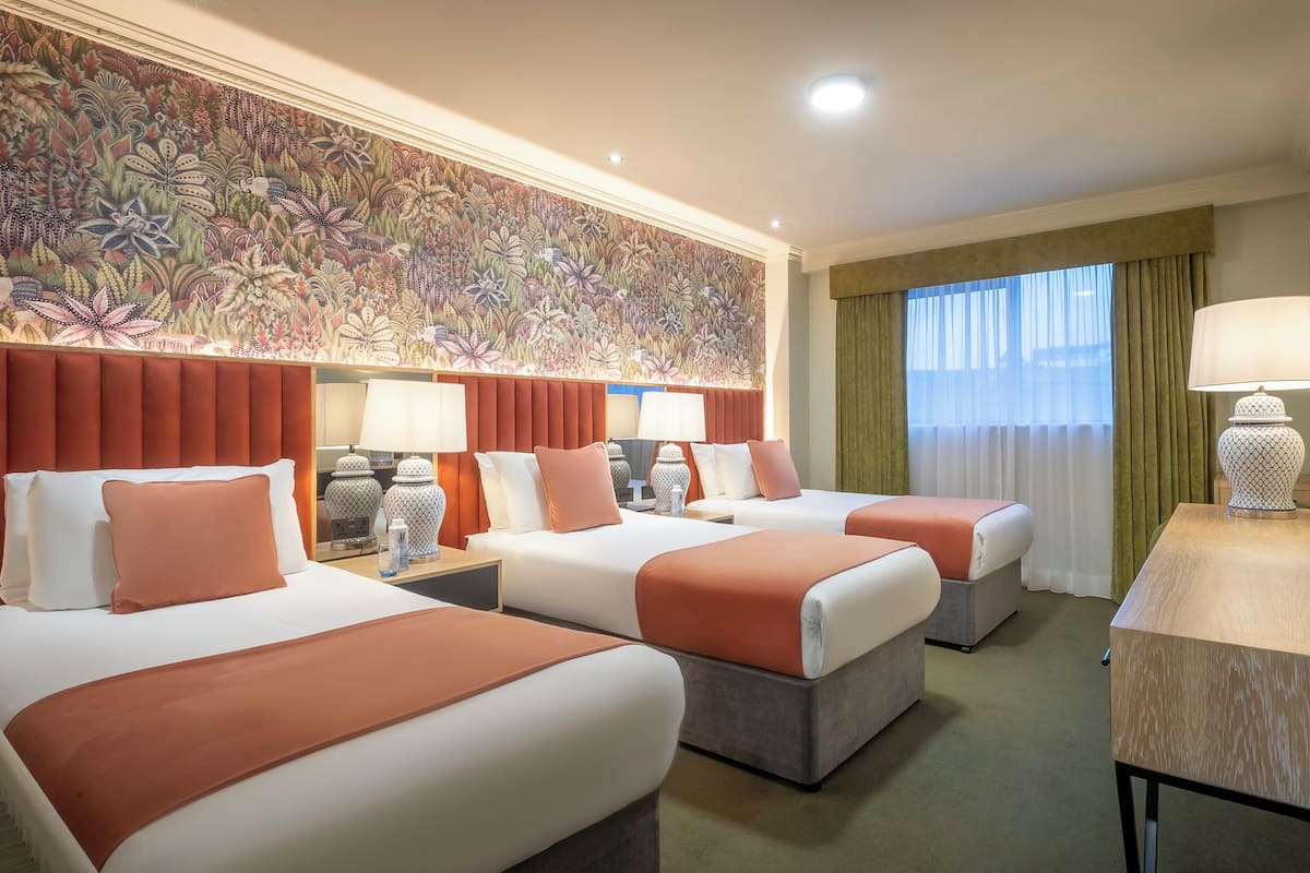 A hotel room with two beds, a large floral-patterned wall, a window with curtains, and a table with a lamp.