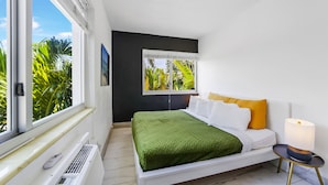 A modern bedroom with a bed, a bedside table with a lamp, a window with a view of greenery, and a radiator. - Harding 8040 Apts By At Mine Hospitality (Miami Beach)