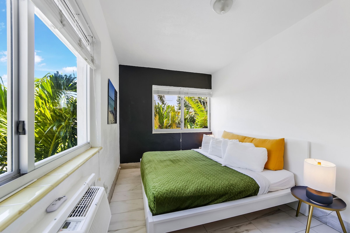 A modern bedroom with a bed, a bedside table with a lamp, a window with a view of greenery, and a radiator.