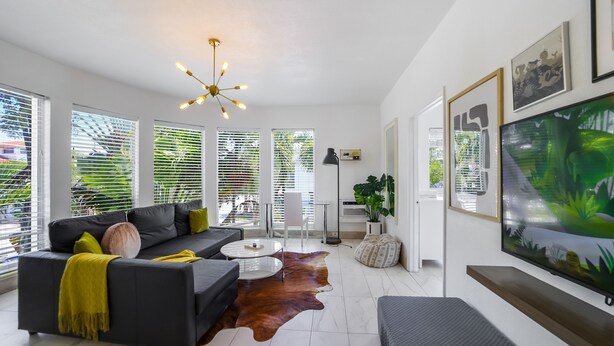 A modern living room with a sofa, coffee table, and a large TV. There are large windows with blinds, a ceiling light, and a plant. - Harding 8040 Apts By At Mine Hospitality (Miami Beach)