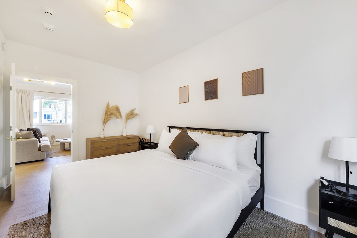 A bedroom with a bed, bedside tables, a dresser, and a door leading to a living area.