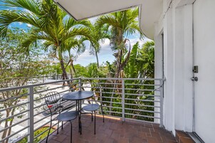 Ein Balkon mit weißer Reling, einem Tisch mit Stühlen und Blick auf Palmen und Gebäude.