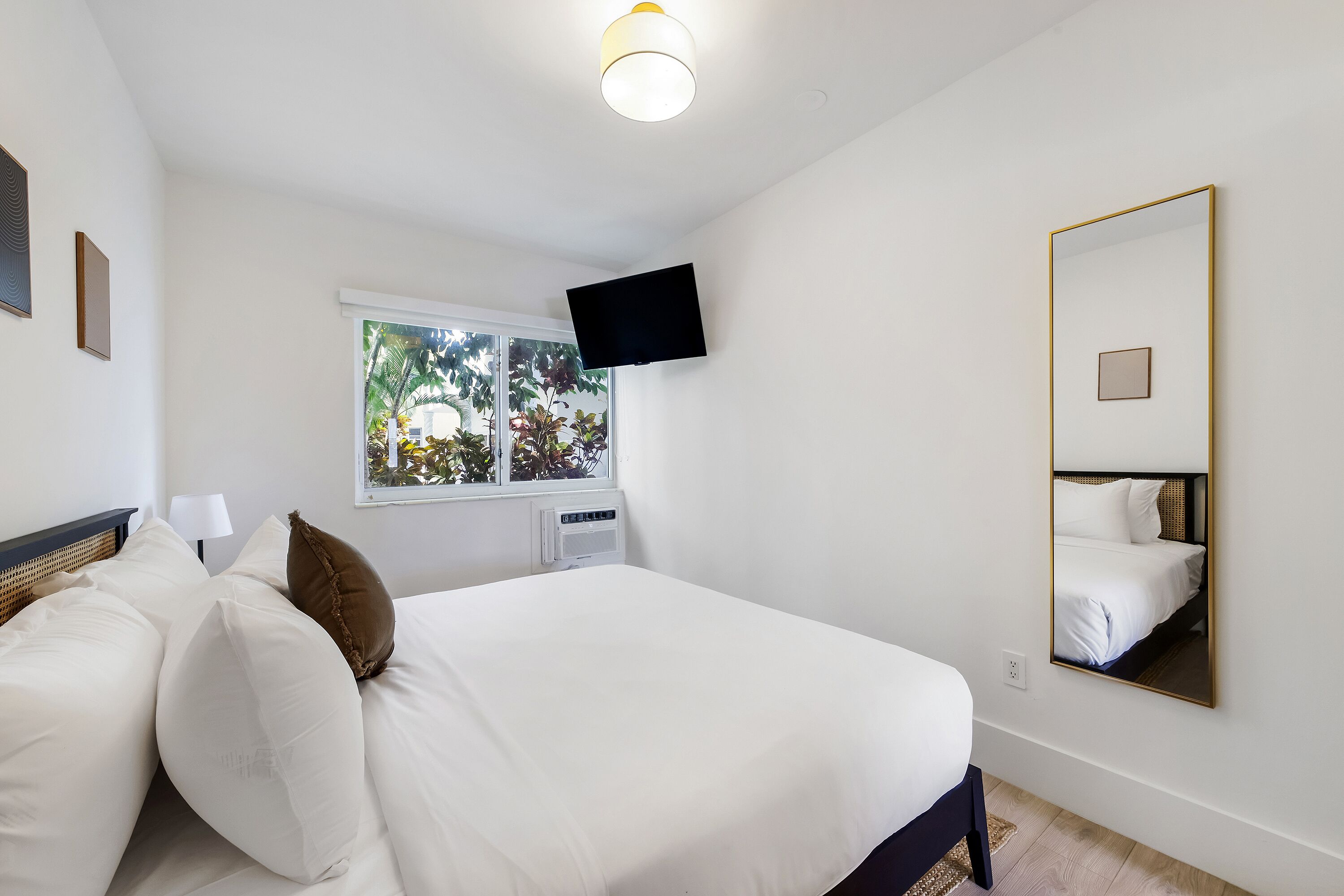 Ein Hotelzimmer mit Bett, Wandfernseher, Fenster mit Blick auf Grünflächen und einem großen Spiegel.
