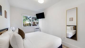 A hotel room with a bed, a TV mounted on the wall, a window with a view of greenery, and a large mirror on the wall.