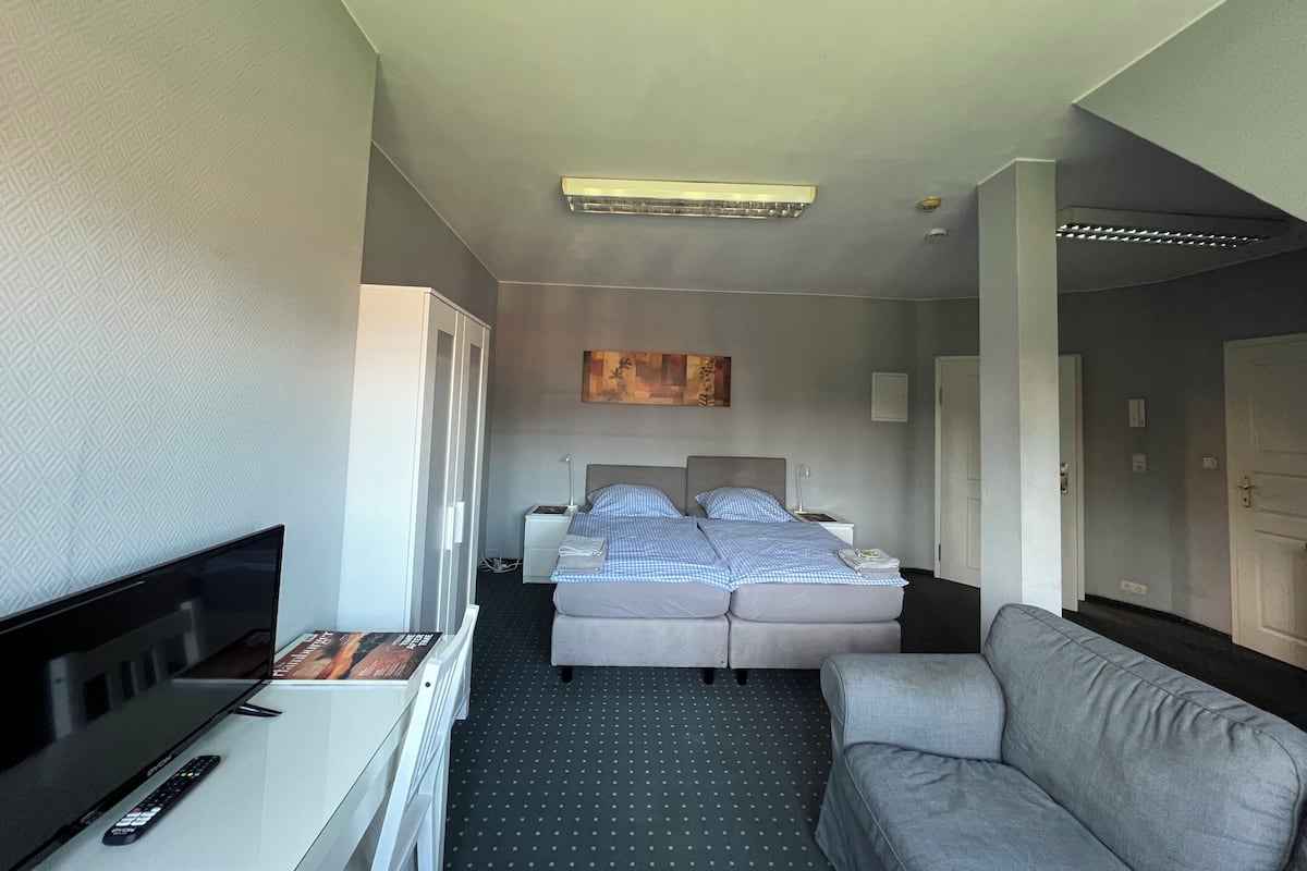 A hotel room with a bed, a TV, a sofa, and a desk with a remote control.