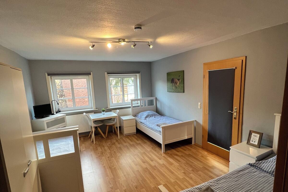 A compact bedroom with a bed, a desk, and a chair. There are two windows providing natural light.