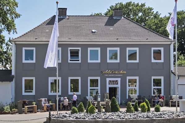 A two-story building with a grey facade and white trim, featuring a sign that reads "Hotel International." There are outdoor seating areas with tables and chairs, and a small garden with shrubs and a fountain. - Bett4you Peiner Hof (Prisdorf)