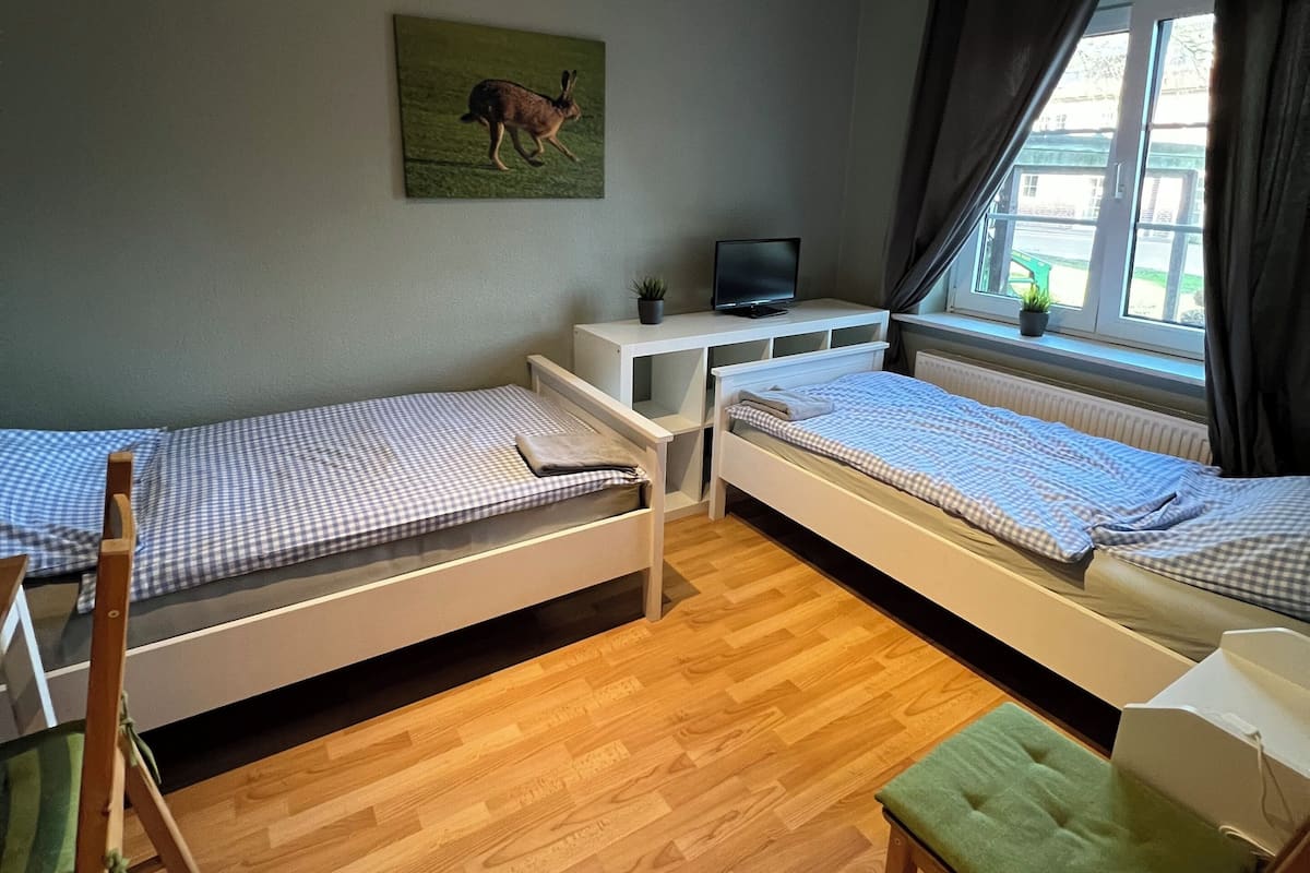 A room with two single beds, a desk with a computer, a chair, a window with curtains, and a painting on the wall.