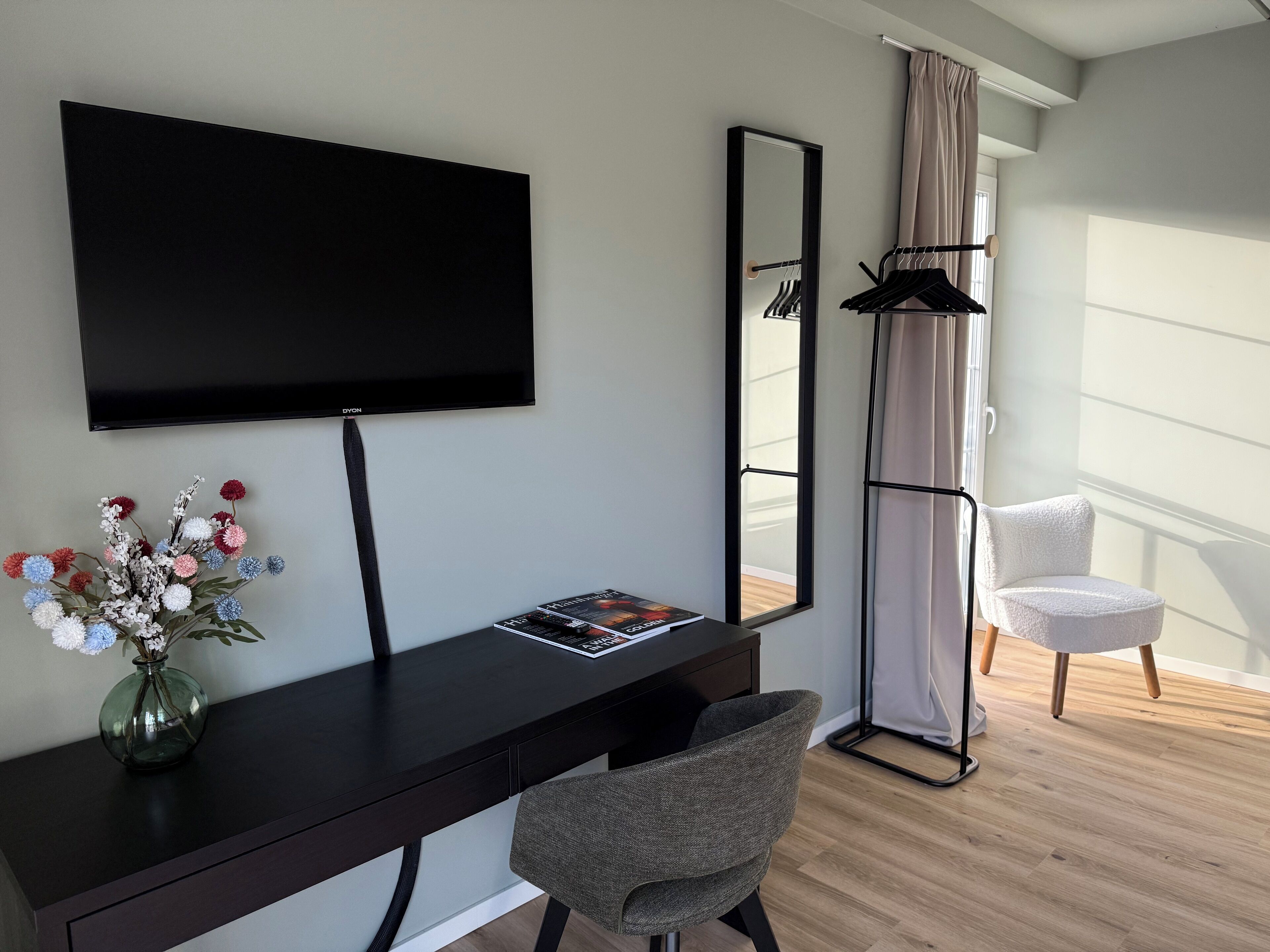Ein modernes Hotelzimmer mit dunklem Holztisch, Flachbildfernseher, Spiegel, Stuhl und einem Fenster mit Vorhängen.