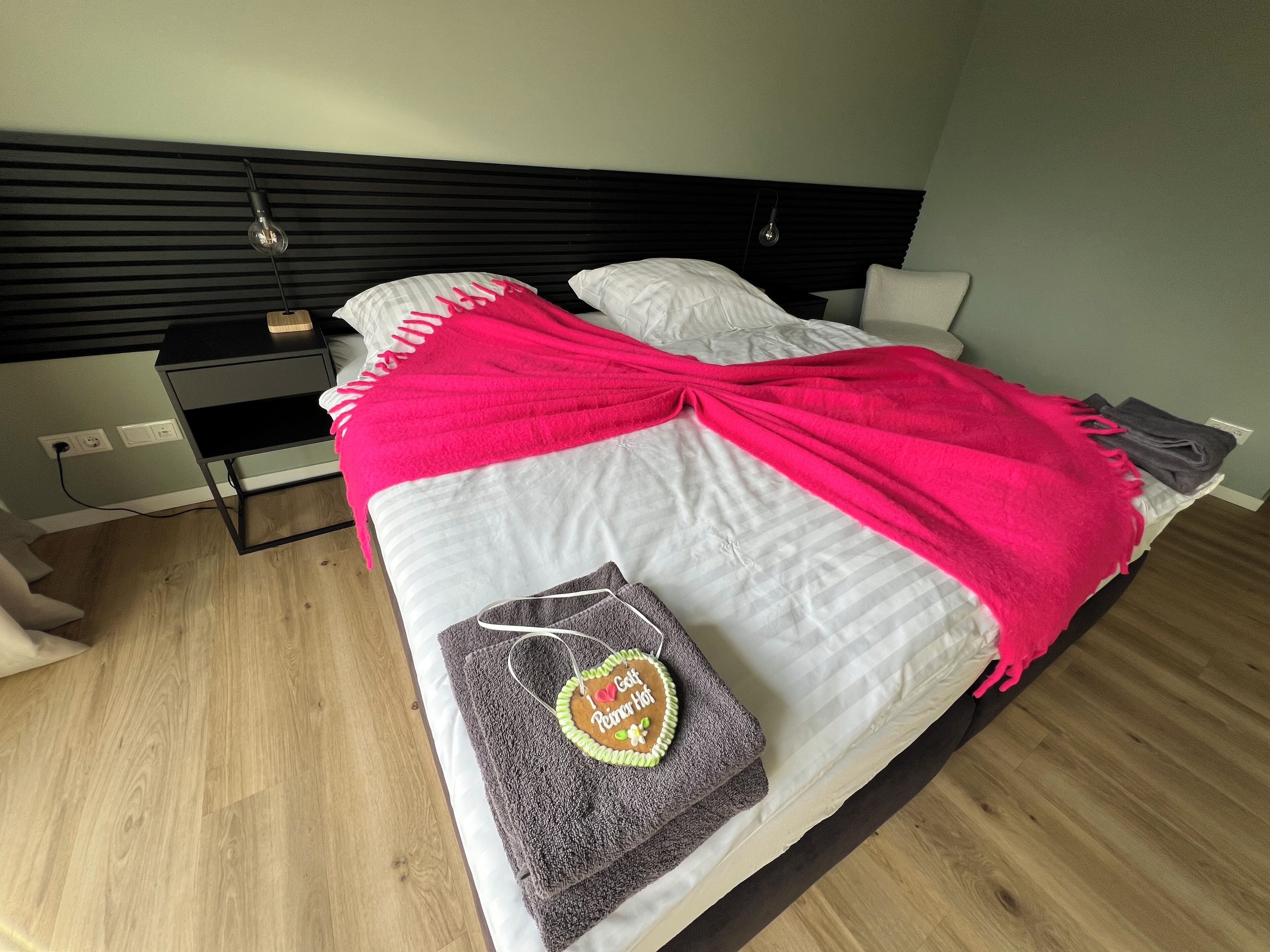 A neatly made bed with a bright pink blanket, a grey towel with a heart design, and a wooden nightstand with a lamp.