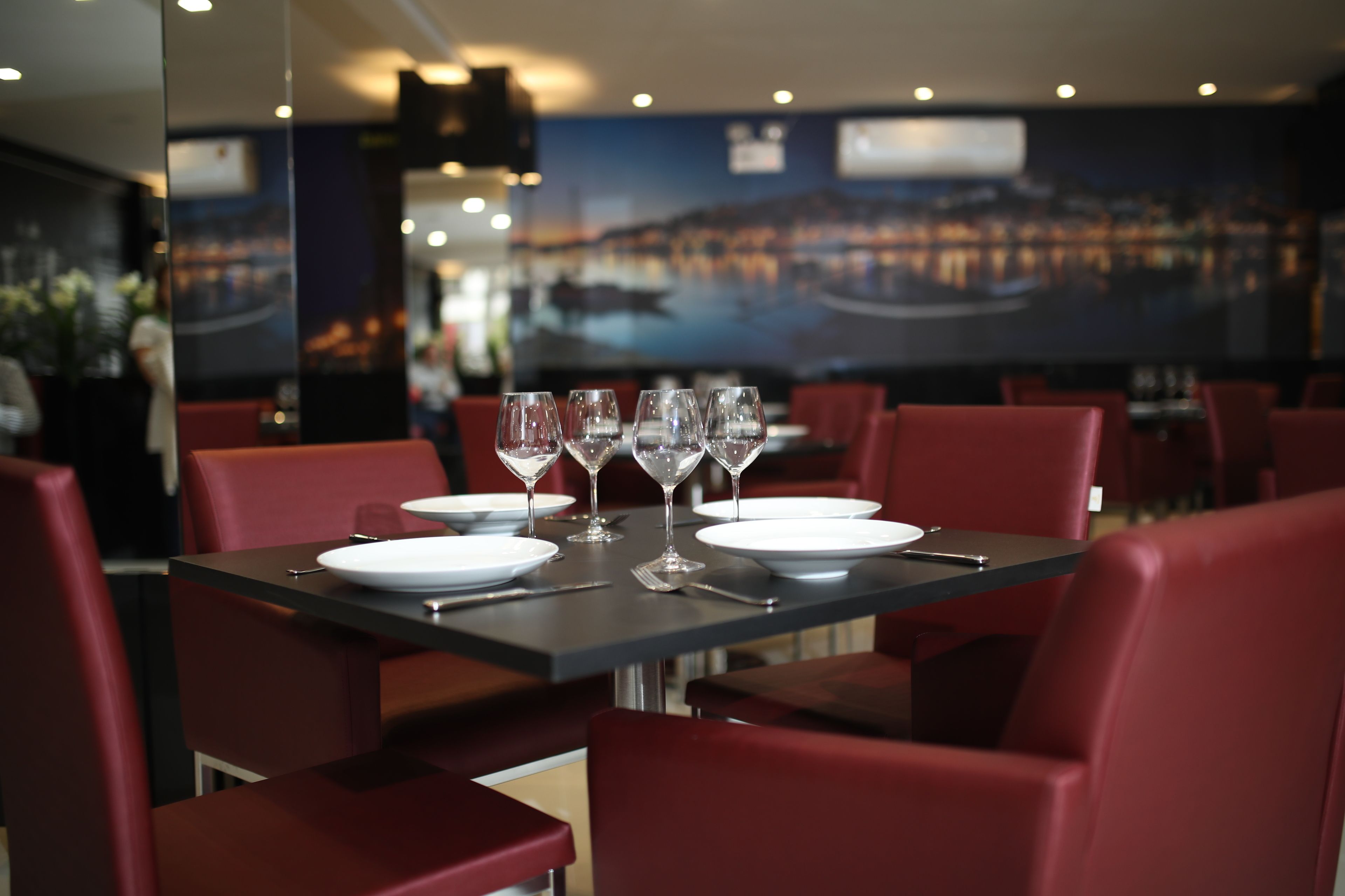 A restaurant with a table set for four, red upholstered chairs, and a cityscape mural on the wall.