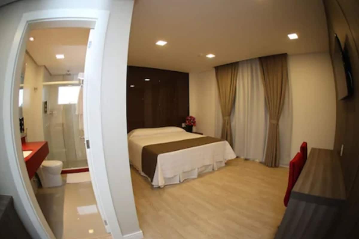 Ein Hotelzimmer mit Bett, Schreibtisch, Stuhl, Duschbereich und einem großen Fenster mit Vorhängen.