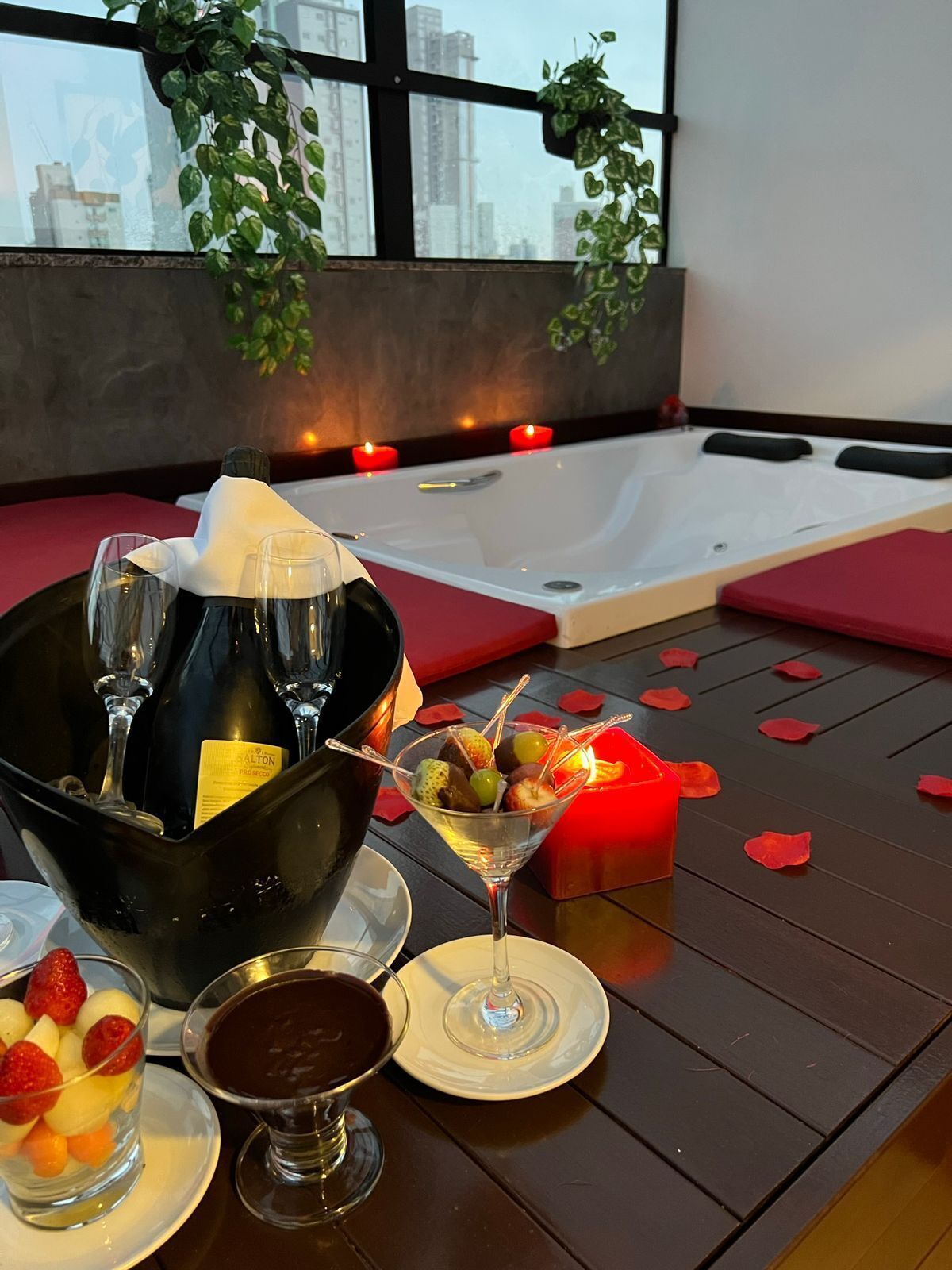 Una habitación con una bañera, una mesa con una botella de champán, una copa de martini, un postre de chocolate y un postre de fruta.
