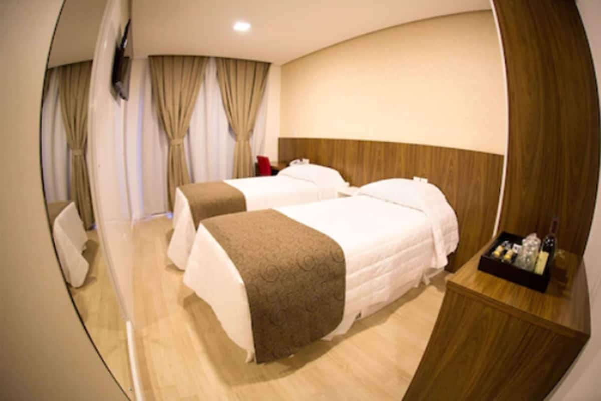 Ein Hotelzimmer mit zwei Betten, einem Holz-Kopfteil, einem Nachttisch mit einer Flasche und Vorhängen.