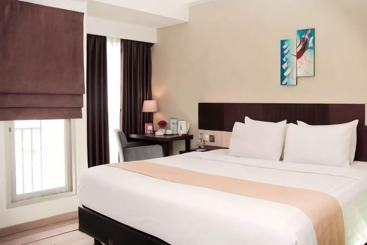 A hotel room with a bed, a desk, a chair, a lamp, and artwork on the wall.