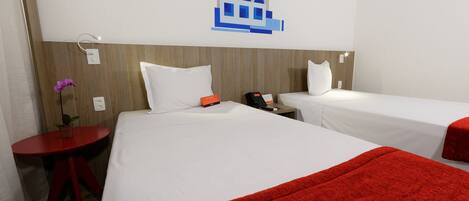 A hotel room with two beds, a red table with a plant, and a wall-mounted light switch.