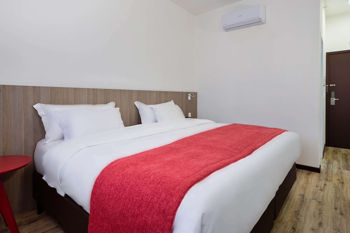Una habitación de hotel con cama, una mesita de noche roja y suelo de madera.