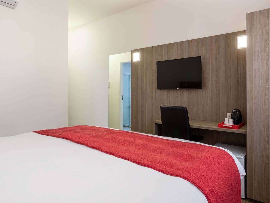 A hotel room with a bed, a desk, a chair, a television, and a small kitchenette area.