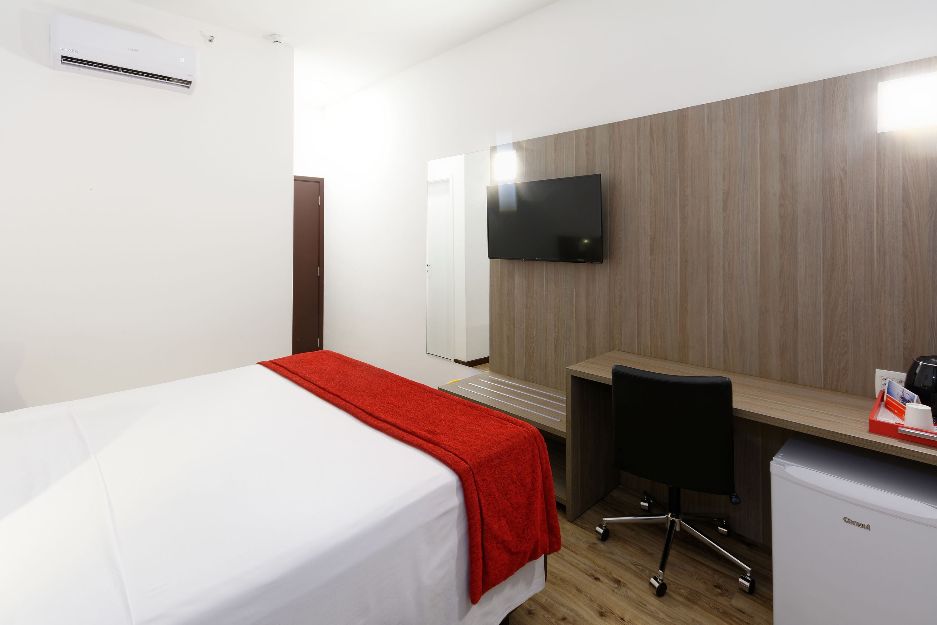 Quarto de hotel com uma cama, escrivaninha, cadeira, televisão fixada na parede e um pequeno frigorífico.