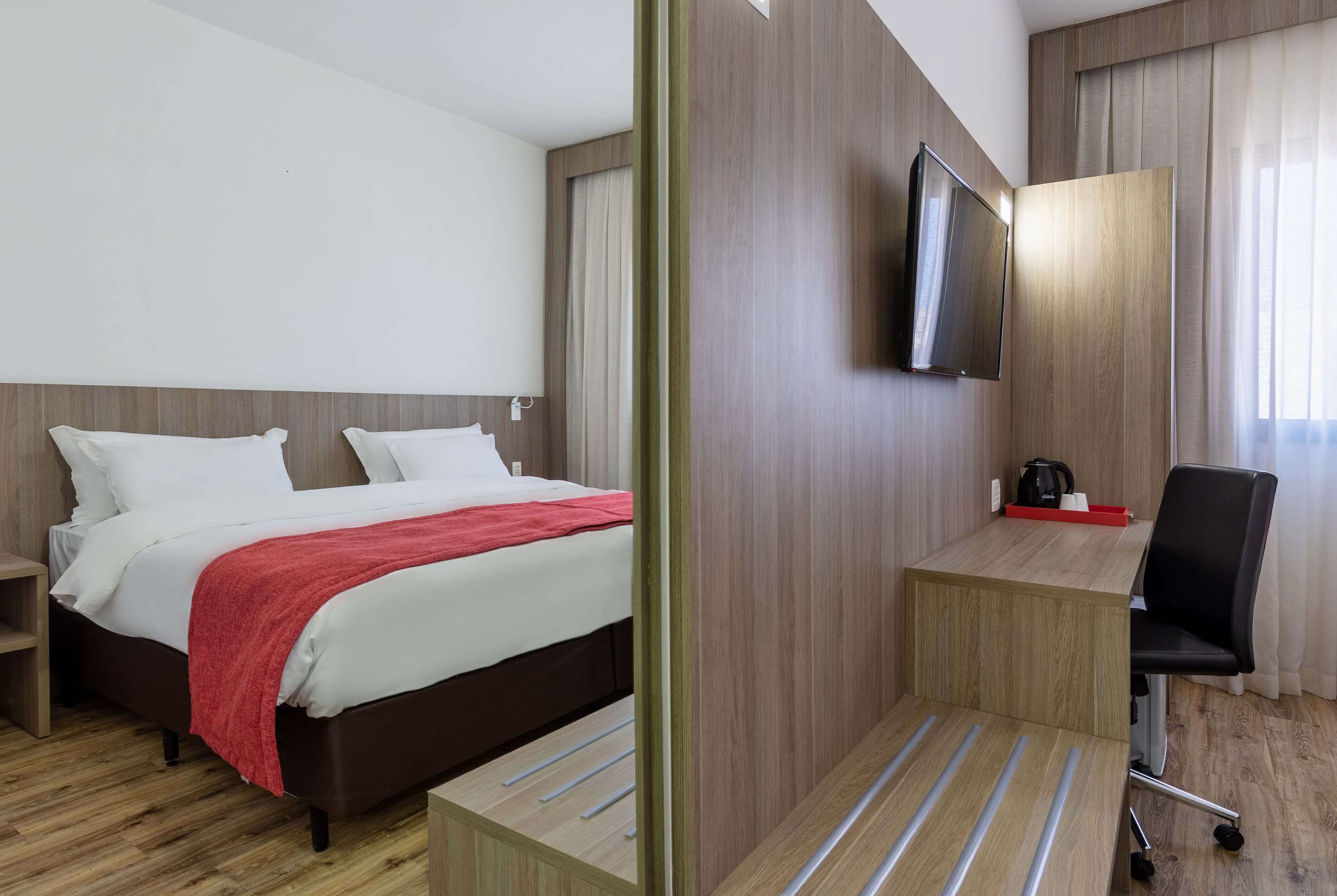A hotel room with a bed, a desk, a chair, a TV, and a window with curtains.