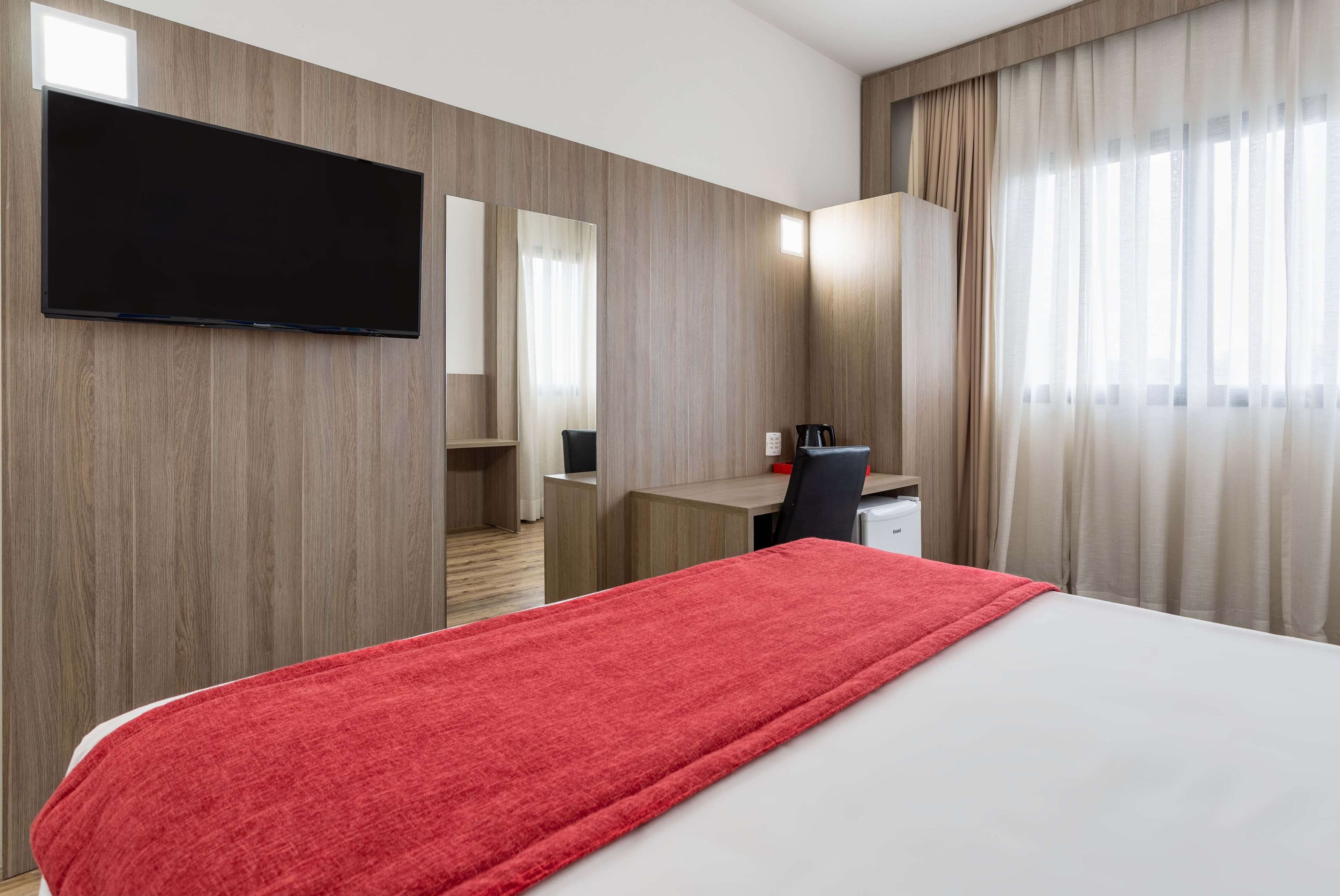 Habitación de hotel con cama, escritorio, silla, televisor y ventana con cortinas.