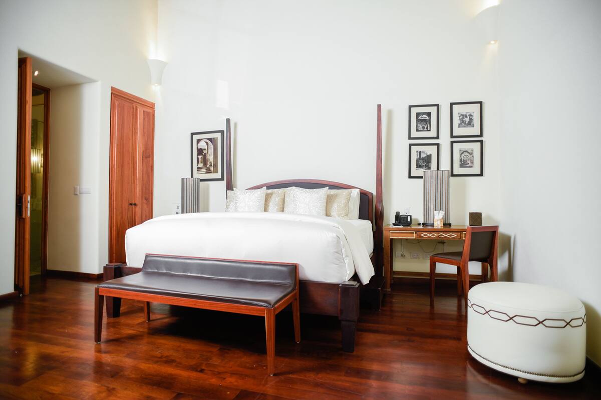 A neatly arranged bedroom with a bed, bench, desk, and chair, along with framed pictures on the wall.