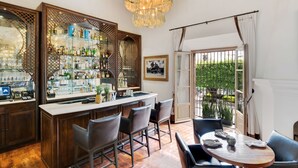 A bar area with a wooden counter, bar stools, and a chandelier. There is a large window with a view of greenery outside. - Hotel Casa Blanca 7 (San Miguel de Allende)