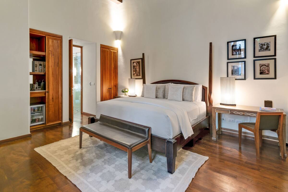 A bedroom with a large bed, wooden wardrobe, bedside table, desk, chair, and a small refrigerator.