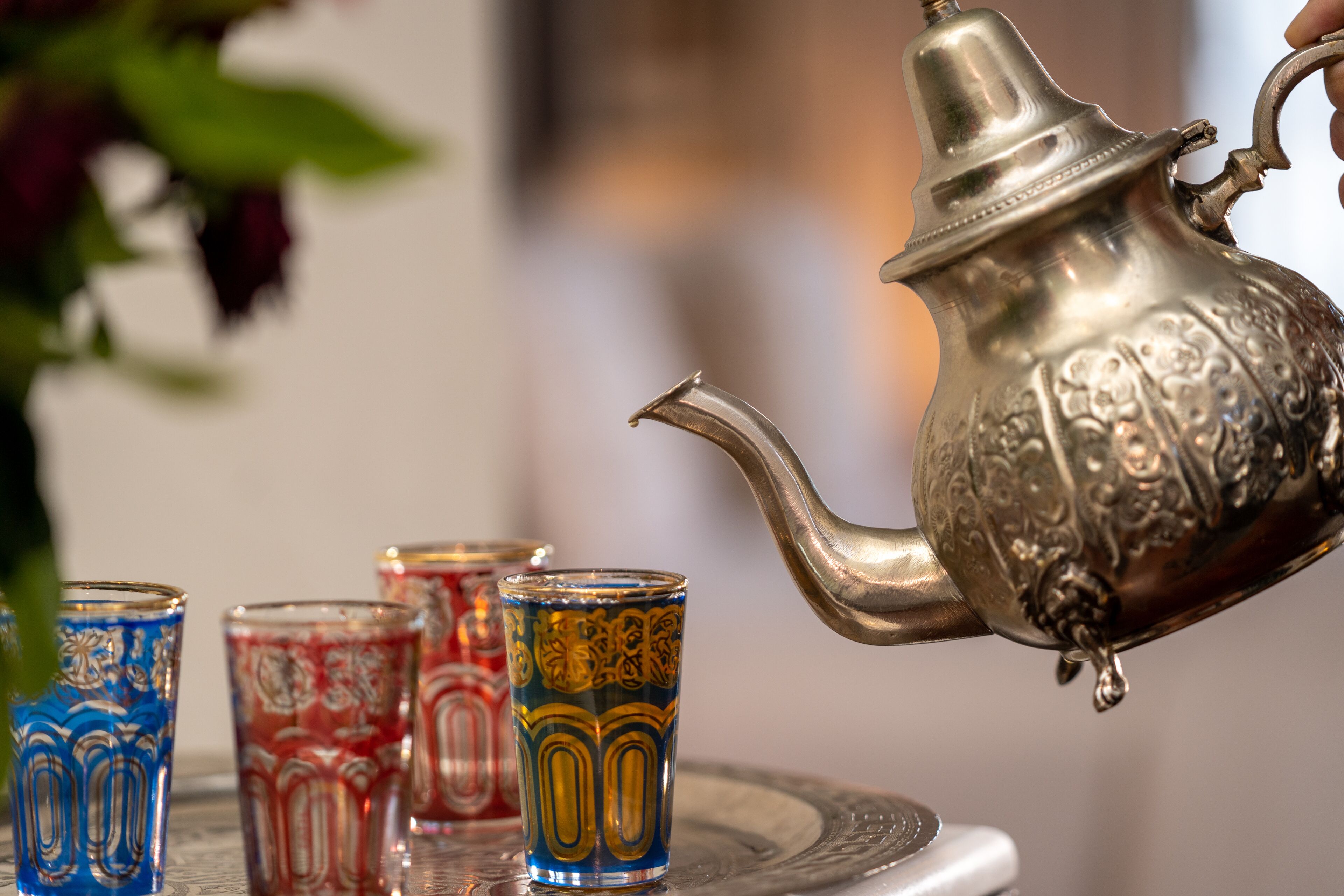 A silver teapot with intricate designs, being poured into a colorful glass.