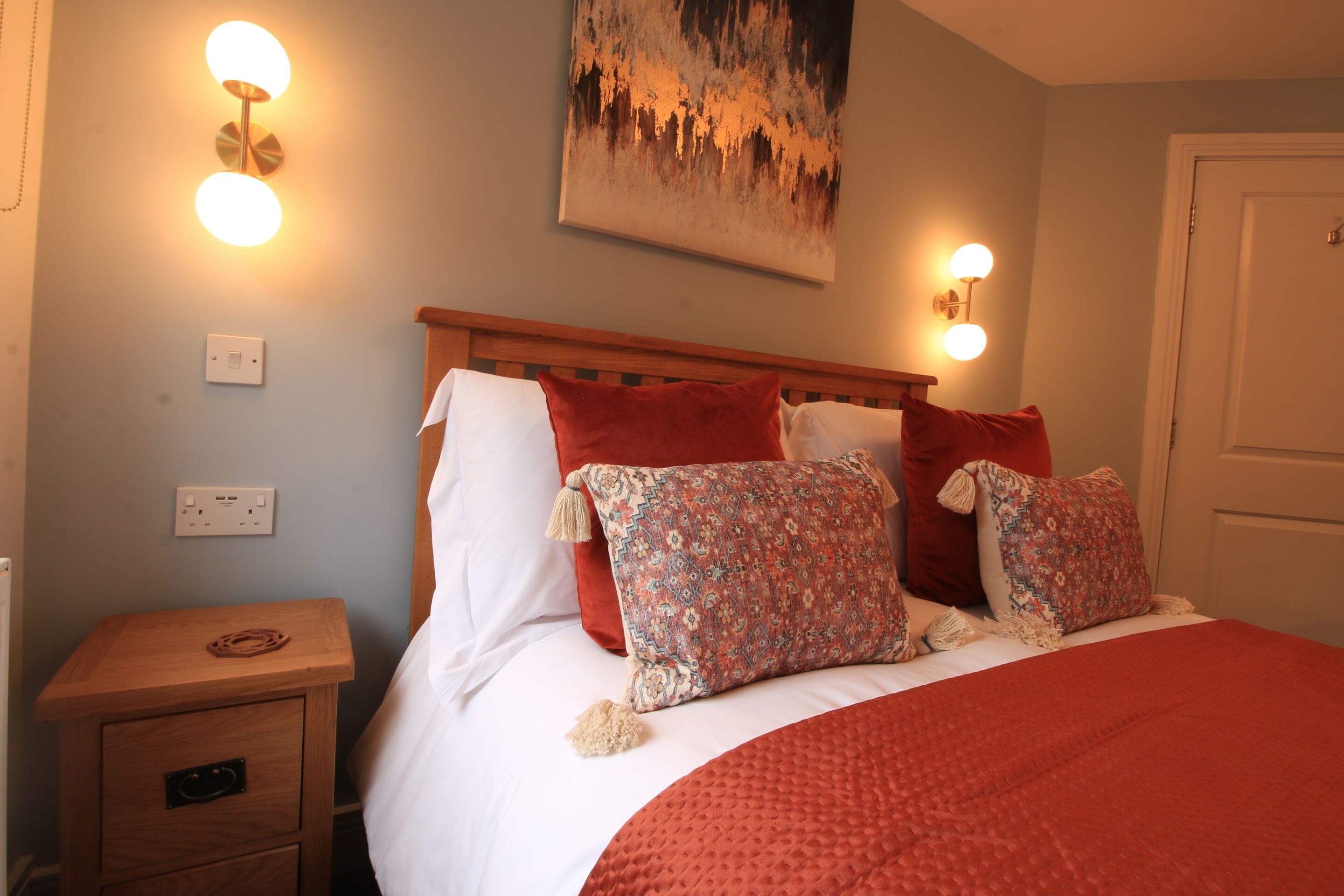 A neatly made bed with a red blanket and patterned pillows, a wooden nightstand with a lamp, and a painting on the wall.