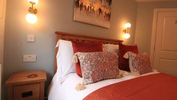 A neatly made bed with a red blanket and patterned pillows, a wooden nightstand with a lamp, and a painting on the wall.