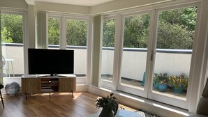 Amplia sala de estar con televisor grande, piso de madera, una mesa redonda y ventanas amplias que ofrecen vistas al exterior.